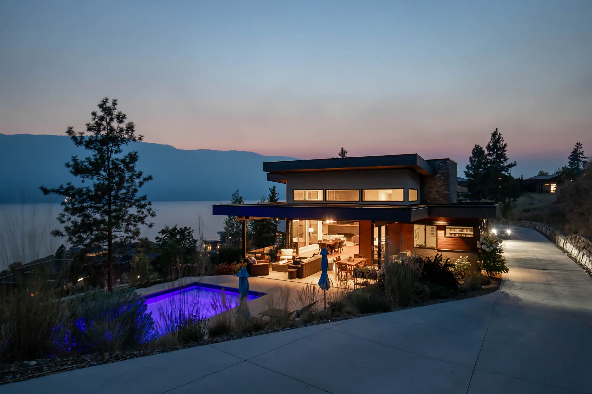 Twilight exterior of a modern Okanagan home with a lit pool and lake view